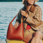 A woman sitting by the water holds a Sienna Large Naomi Shoulder Bag. She wears a plaid shirt, shorts, and a wide-brim hat, with the blue lake in the background.