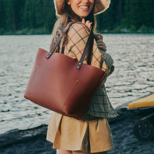 A woman outdoors by the water wearing a wide-brimmed hat and carrying a large brown leather tote bag over her shoulder.