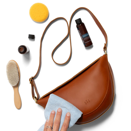 All Variants | Person using a soft towel to rub in Revitalizing Distressed Leather Conditioner into a purse surrounded by a brush, sponge, and containers of the conditioner.