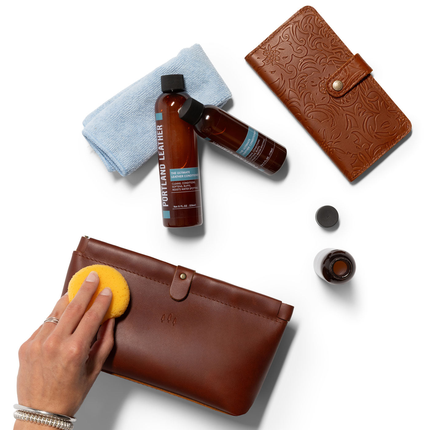 All Variants | A hand is applying the Ultimate Leather Conditioner to a leather pouch while the 4oz and 8oz lay on a towel